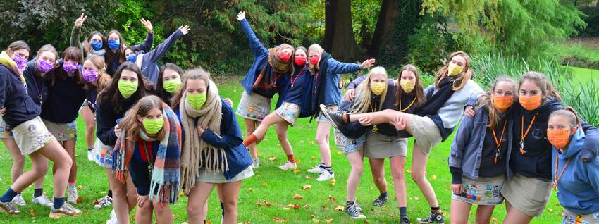 Hier zie je een prachtige groepsfoto van de meisjesleiding Duffel-Oost. Ze staan in groepjes per afdeling en dragen een mondmasker in hun afdelingskleur.